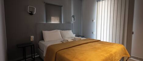 Un dormitorio con cama, mesita de noche, lámpara de pared y ventana con persianas.