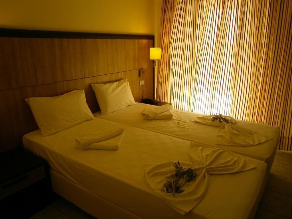 A hotel room with a large bed, a wooden headboard, a nightstand, and a window with vertical blinds.