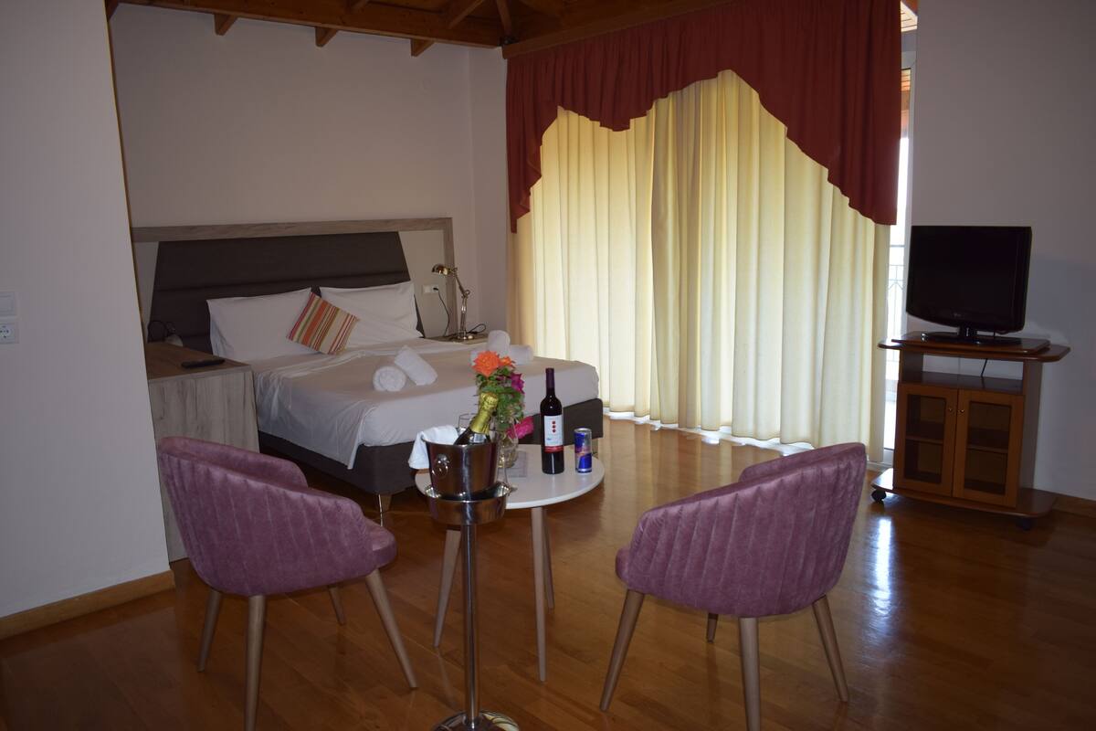 A hotel room with a bed, a TV, a table with a bottle and flowers, and two chairs.