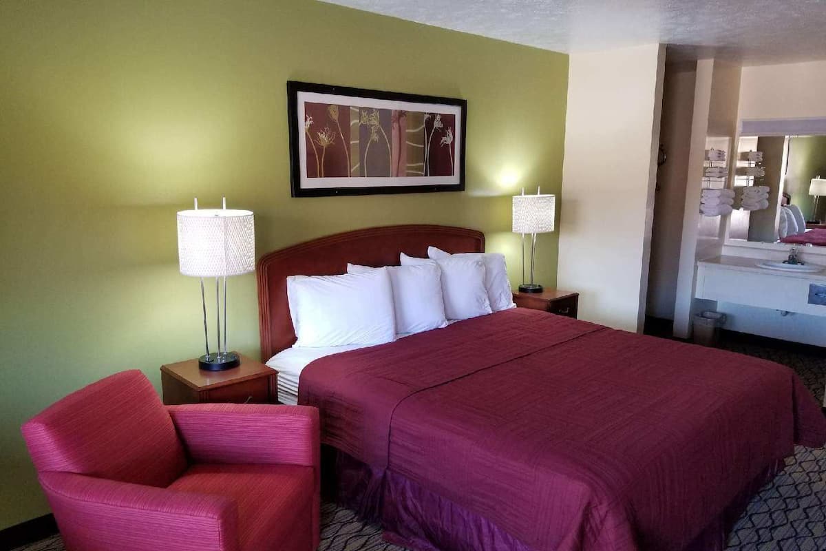 Ein Hotelzimmer mit einem großen Bett, einem roten Sessel, einem Nachttisch mit Lampe und einem gerahmten Bild an der Wand.