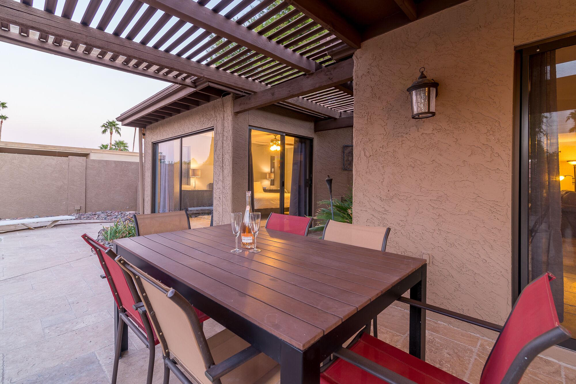An outdoor patio with a wooden table and red chairs, a pergola overhead, and a view into a room with a bed and a lamp.