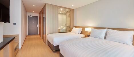 Une chambre d’hôtel avec deux lits, un plancher en bois, un téléviseur fixé au mur et une salle de bain visible à travers une porte en verre.
