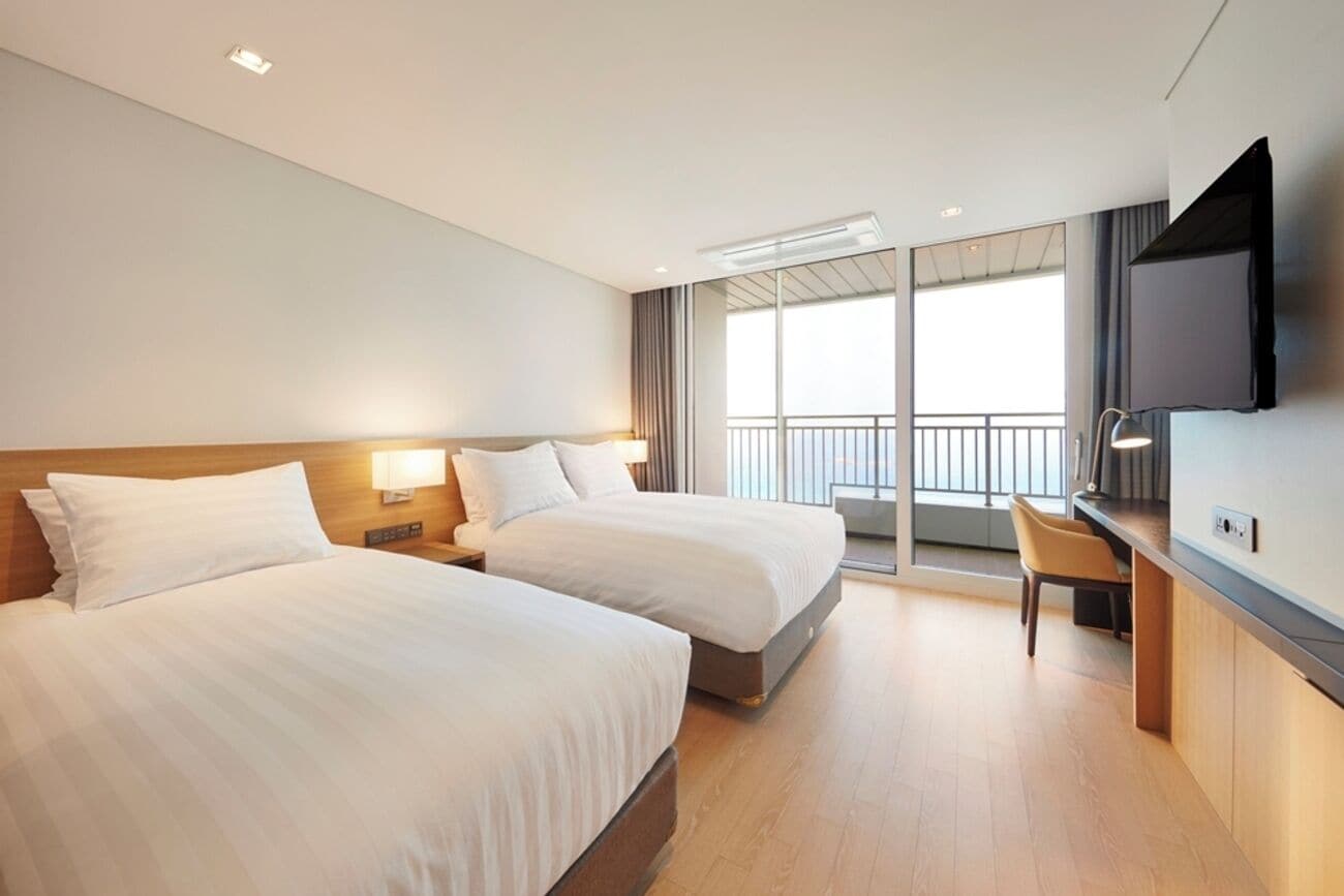 A hotel room with two beds, a desk, a TV, and a balcony with a view.