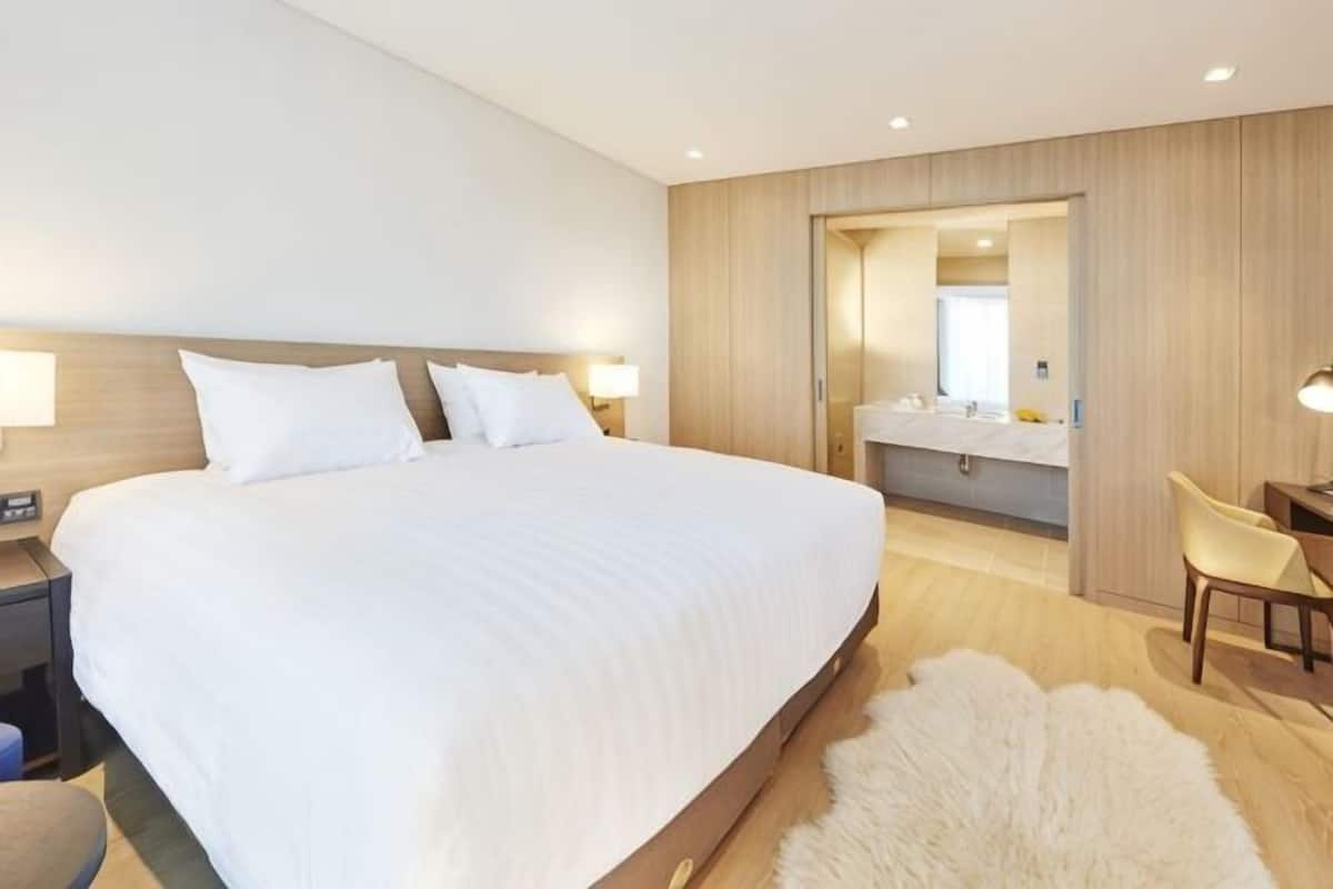 A modern hotel room with a large bed, a desk, a chair, a mirror, and a bathroom visible through an open door.