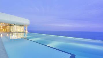 Una piscina infinita moderna con vista despejada al océano, un edificio blanco con grandes ventanales y un cielo vespertino tranquilo.