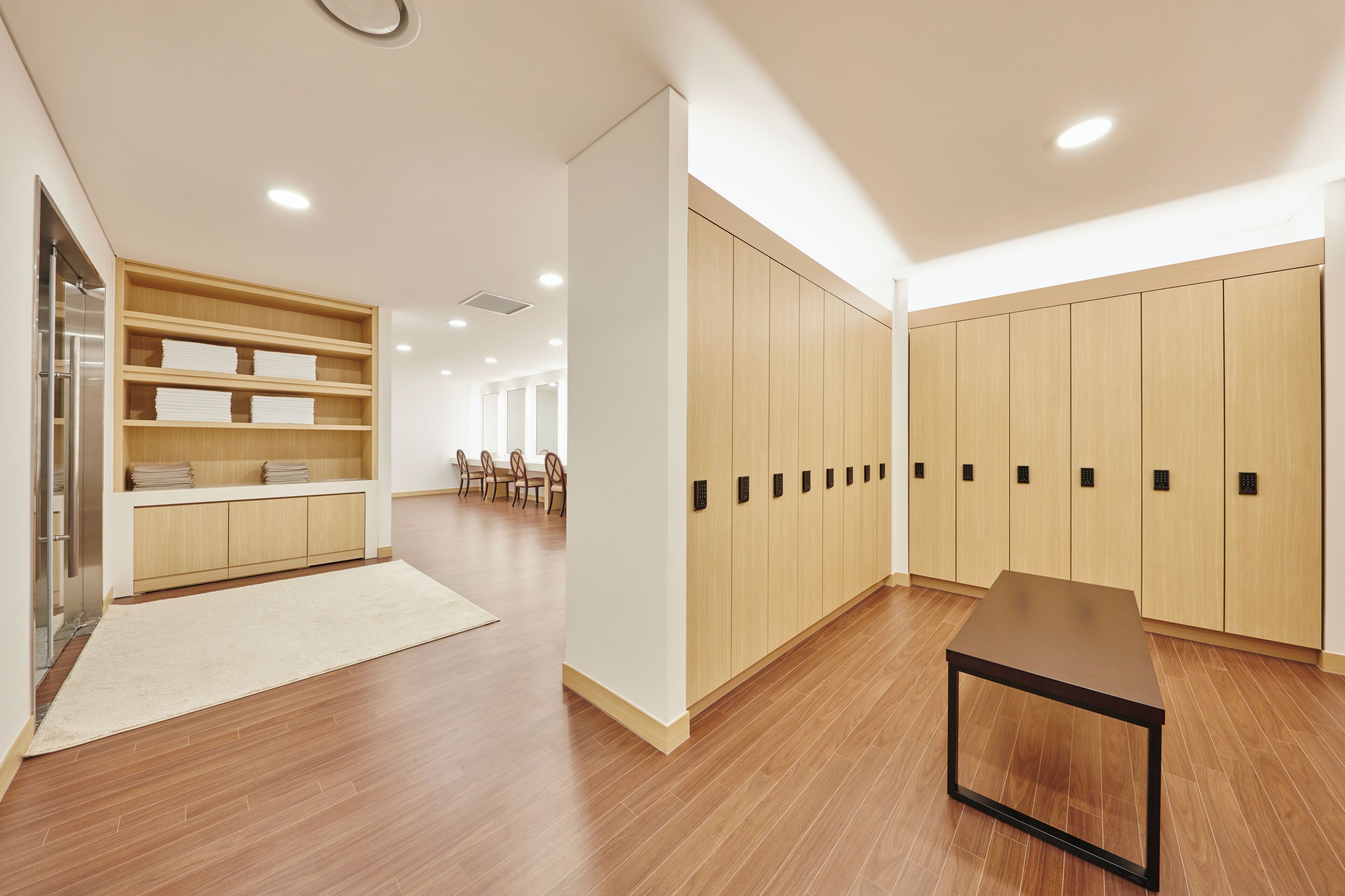 A modern, well-lit room with wooden flooring, a beige rug, a table, a seating area with chairs, and a wardrobe with multiple doors.