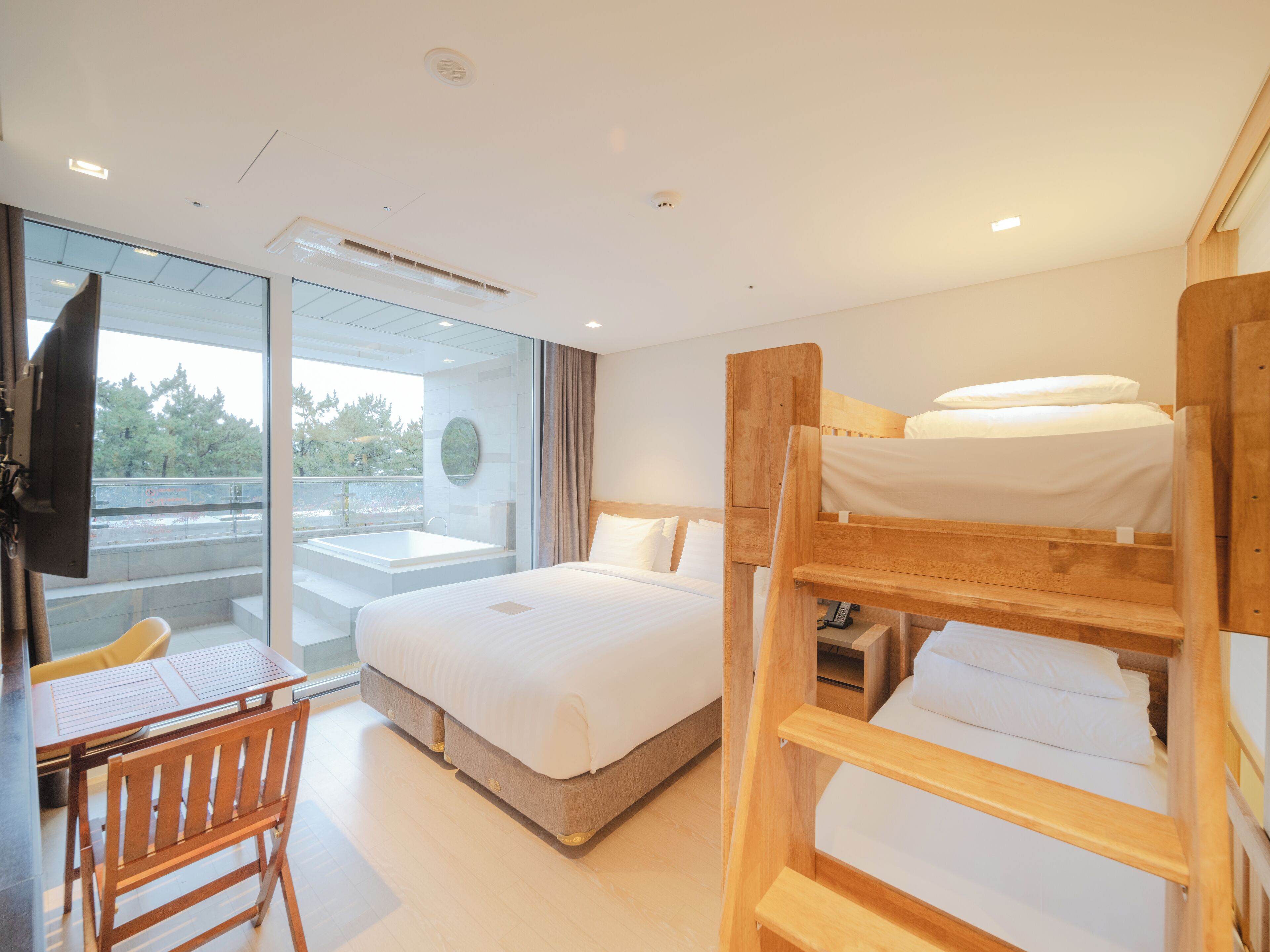 A modern hotel room with a large bed, a wooden bunk bed, a desk with a chair, and a glass wall offering a view of the outdoors.