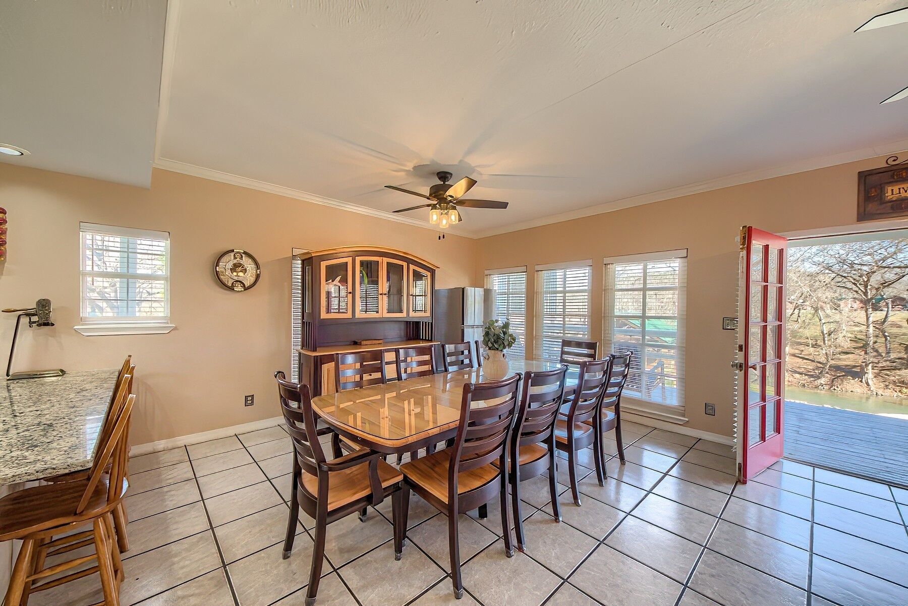A dining area with a large wooden table and chairs, a ceiling fan, and a view of the outdoors through large windows.
