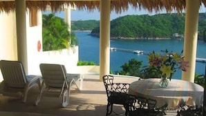 A covered outdoor seating area with chairs and a table, overlooking a coastal landscape with greenery and a pier. - Villa Punta Sta Cruz 3BR (Santa María Huatulco)