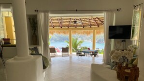 A spacious living area with a large window overlooking a scenic ocean view, a dining table with chairs, and a television mounted on the wall. - Villa Punta Sta Cruz 3BR (Santa María Huatulco)
