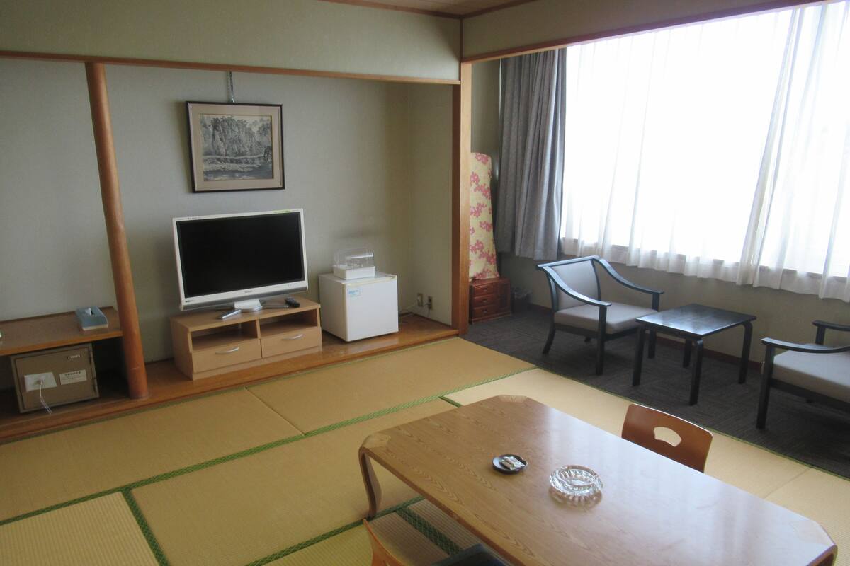A traditional Japanese room with tatami flooring, a television, a small refrigerator, a desk, and a window with curtains.