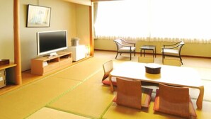 A traditional Japanese-style room with tatami flooring, a low table with cushions, a television, and a window with curtains.
