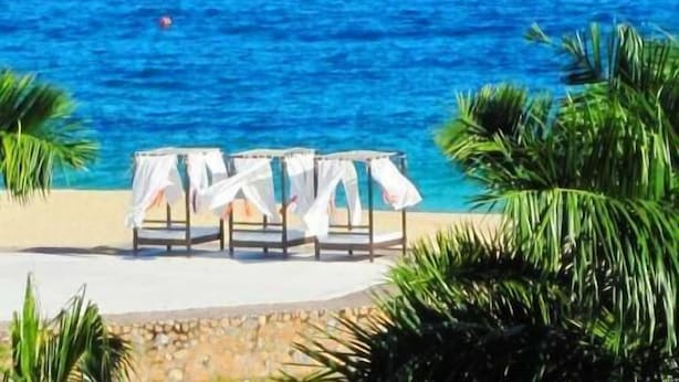 A beachside setting with a row of white-covered chairs and tables, palm trees, and a clear blue sea. - Condominio SDM Chahue 2BR (Santa María Huatulco)