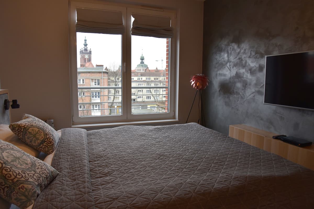 A bedroom with a large bed, a TV on the wall, and a window with a city view.