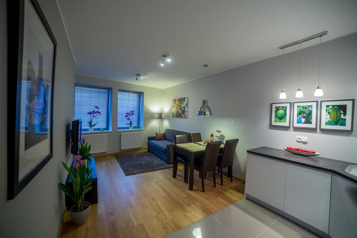 A modern living room with a sofa, dining table, and chairs, a kitchen area with a counter and cabinets, and a hallway with framed pictures and a potted plant.