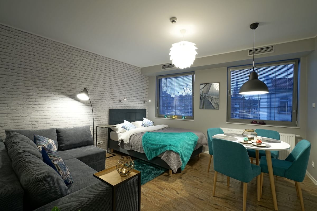 A modern bedroom with a brick wall, a bed with a teal blanket, a grey sofa, a dining table with chairs, and large windows with blinds.