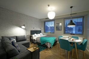 Ein modernes Schlafzimmer mit Backsteinwand, einem Bett mit türkisfarbener Decke, einem grauen Sofa, einem Esstisch mit Stühlen und großen Fenstern mit Jalousien.