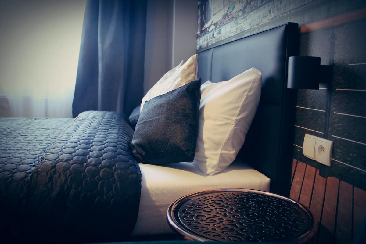 A neatly made bed with a patterned bedspread and pillows, a round side table with a decorative top, and a wall-mounted light fixture.