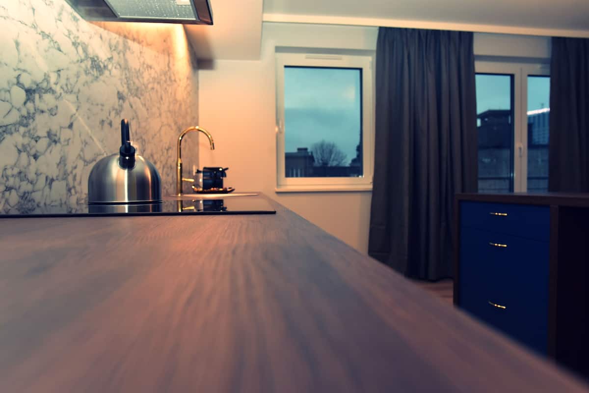 A modern kitchen with a marble backsplash, a stainless steel kettle, and a window with curtains.