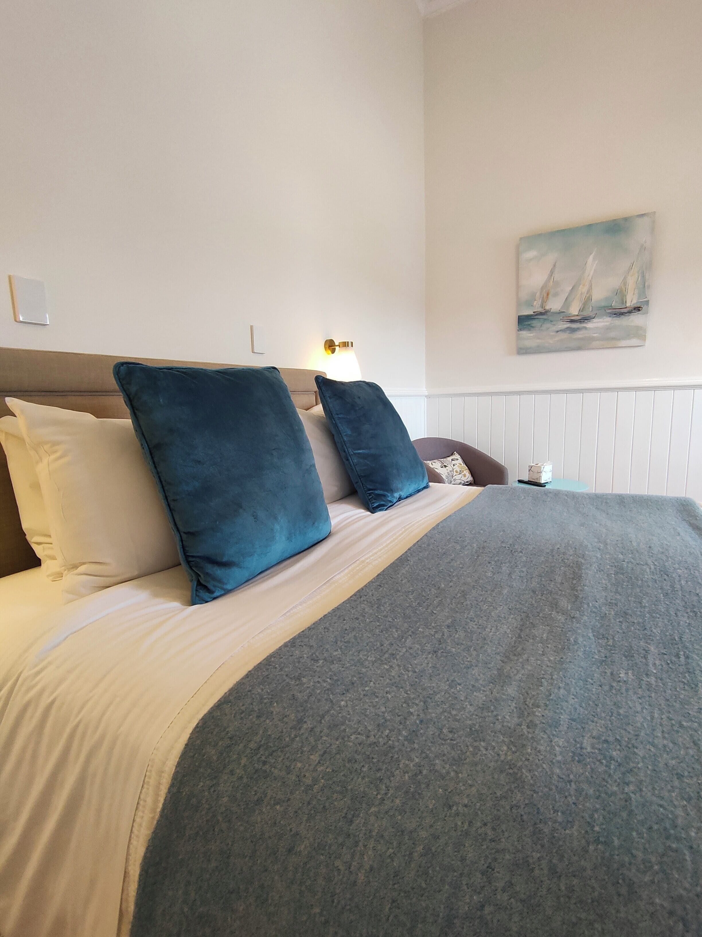 A neatly made bed with a grey blanket and two blue pillows, a wall-mounted lamp, and a painting of sailboats.