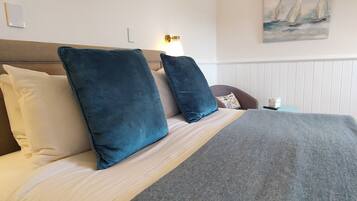 A neatly made bed with a grey blanket and two blue pillows, a wall-mounted lamp, and a painting of sailboats.