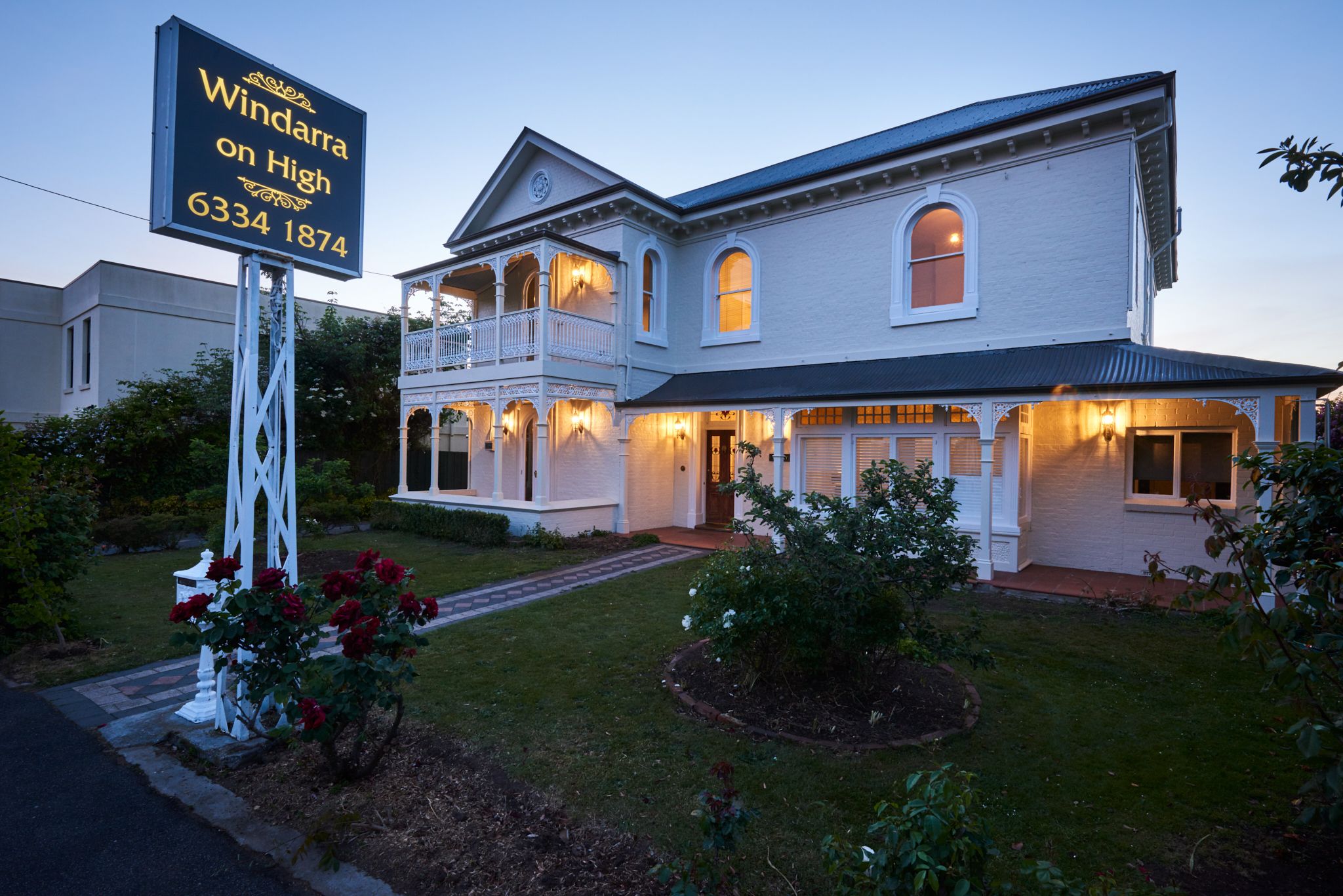 A large, two-story building with a balcony and multiple windows, surrounded by a well-maintained lawn and flowering plants.