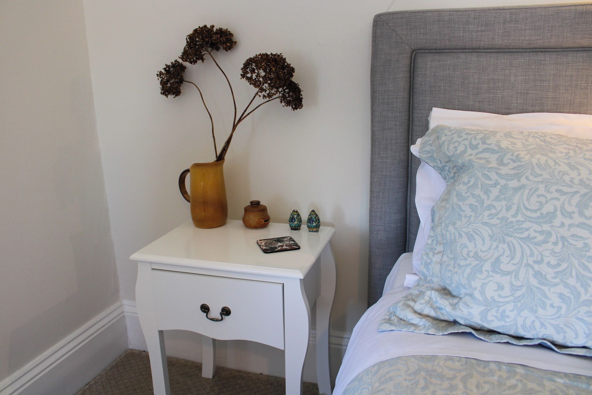 Ein Schlafzimmer mit einem weißen Nachttisch, einer Vase mit getrockneten Blumen, einem kleinen Dekoartikel und einem gemusterten Kissen auf einem Bett mit grauem Kopfteil.