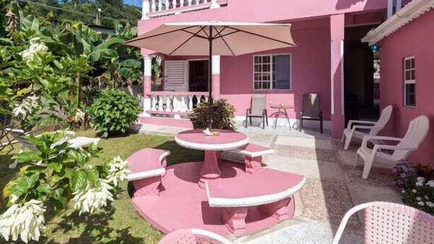 A pink building with a balcony, outdoor seating area with tables and chairs, and a garden with various plants. - Baywatch Apartments (Castara)
