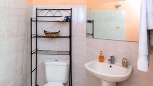 A bathroom with a toilet, a sink, a towel rack, and a shelf with toiletries. - Baywatch Apartments (Castara)