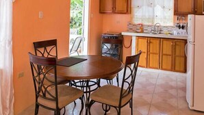 A kitchen with a round table and four chairs, wooden cabinets, a refrigerator, and a window with a view of greenery. - Baywatch Apartments (Castara)