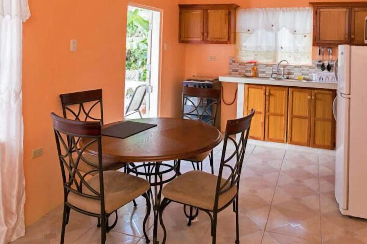 A kitchen with a round table and four chairs, wooden cabinets, a refrigerator, and a window with a view of greenery.