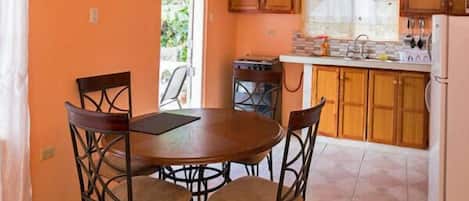 A kitchen with a round table and four chairs, wooden cabinets, a refrigerator, and a window with a view of greenery.