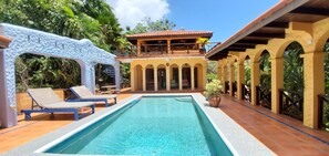 A swimming pool with lounge chairs, a covered patio area, and a building with arched columns.