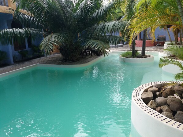 Una piscina con agua cristalina, rodeada de palmeras y un pequeño jardín con rocas.