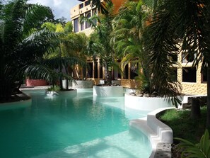 Uma área de piscina com água cristalina, rodeada por palmeiras e um edifício com fachada texturizada.