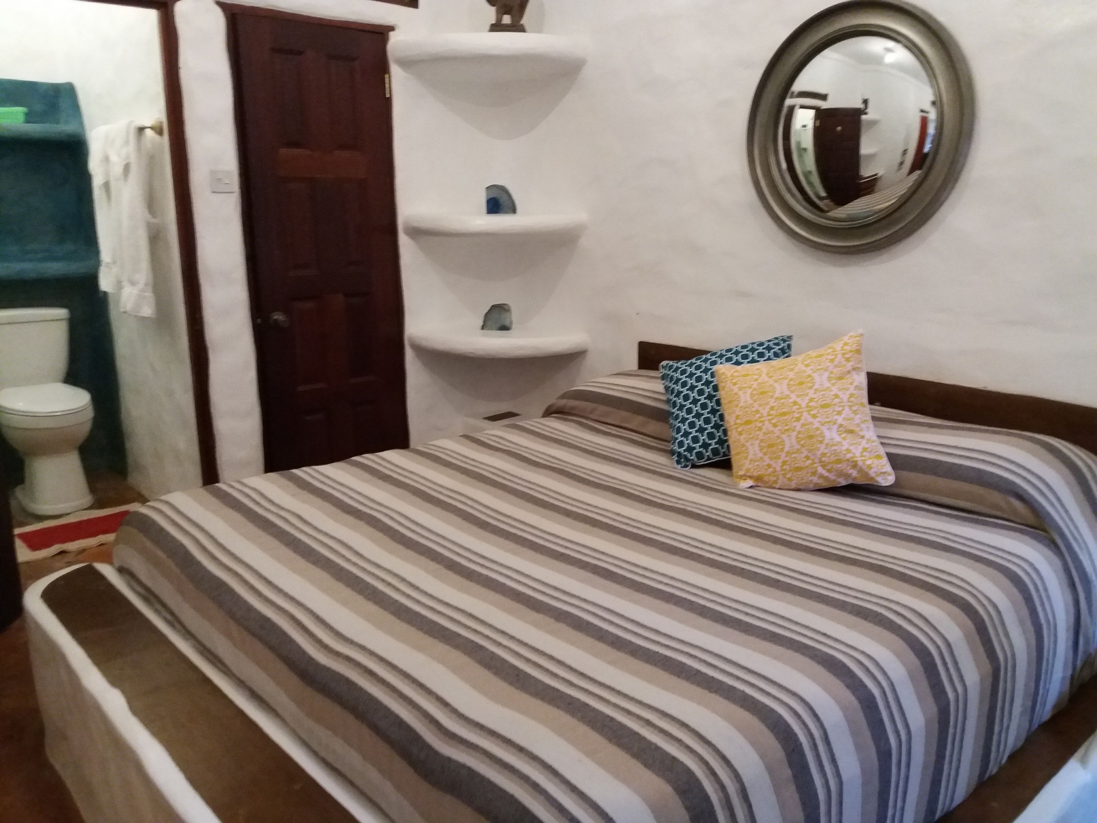 Un dormitorio con colcha a rayas, cabecera de madera, espejo y estante blanco con adornos.