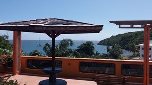 A patio with a wooden umbrella, a bench, and a view of the sea and a hill.