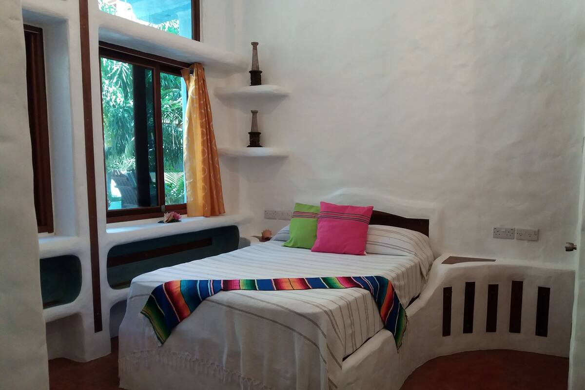 A bedroom with a bed, a window with curtains, a small shelf, and a power outlet on the wall.
