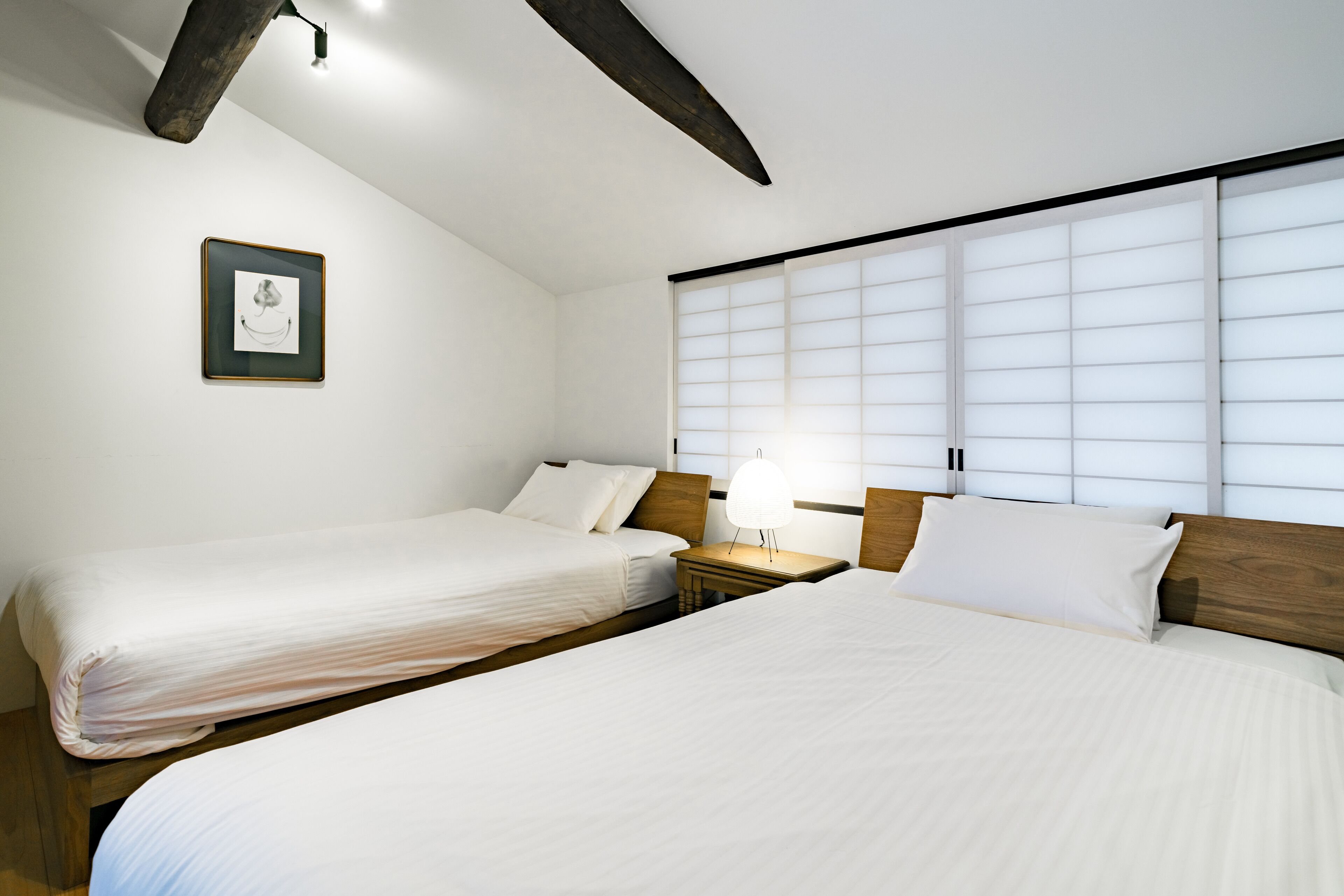 A hotel room with two beds, a nightstand with a lamp, a framed picture on the wall, and a large window with blinds.