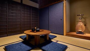 A tatami mat room with a round table, cushions, a tea set, and a traditional Japanese instrument.