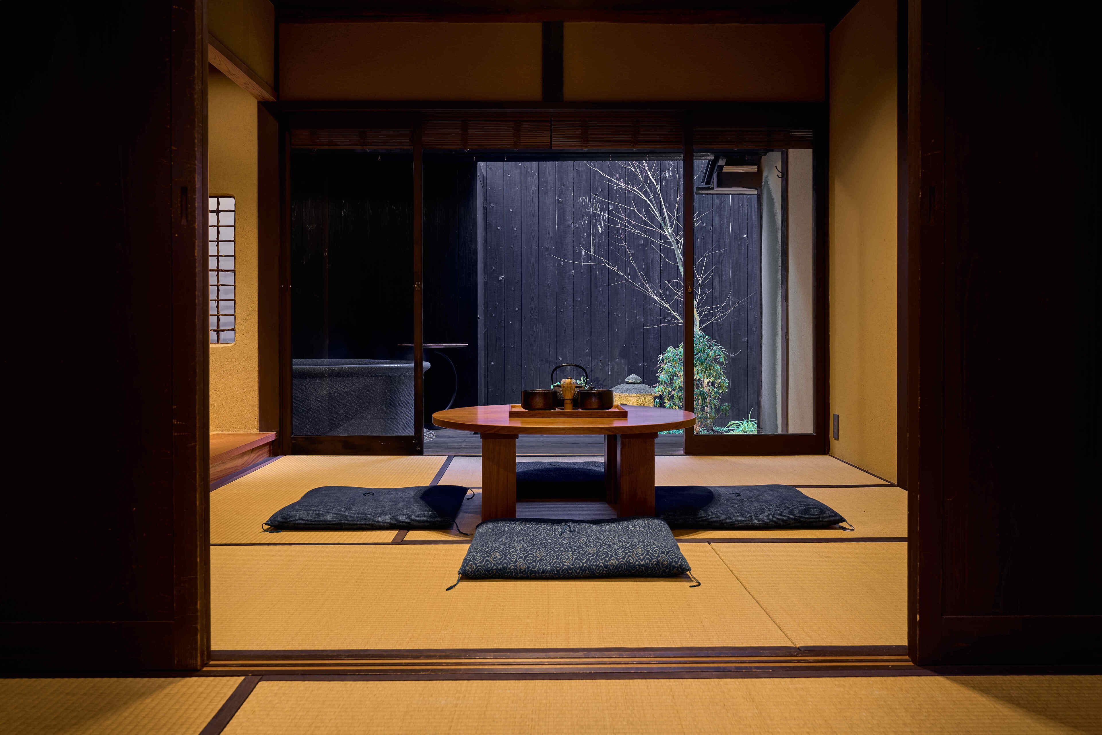 Une pièce de style japonais traditionnel, avec un sol recouvert de tatamis, une petite table sur laquelle est disposé un service à thé, et une vue sur un arbre à travers une porte coulissante.