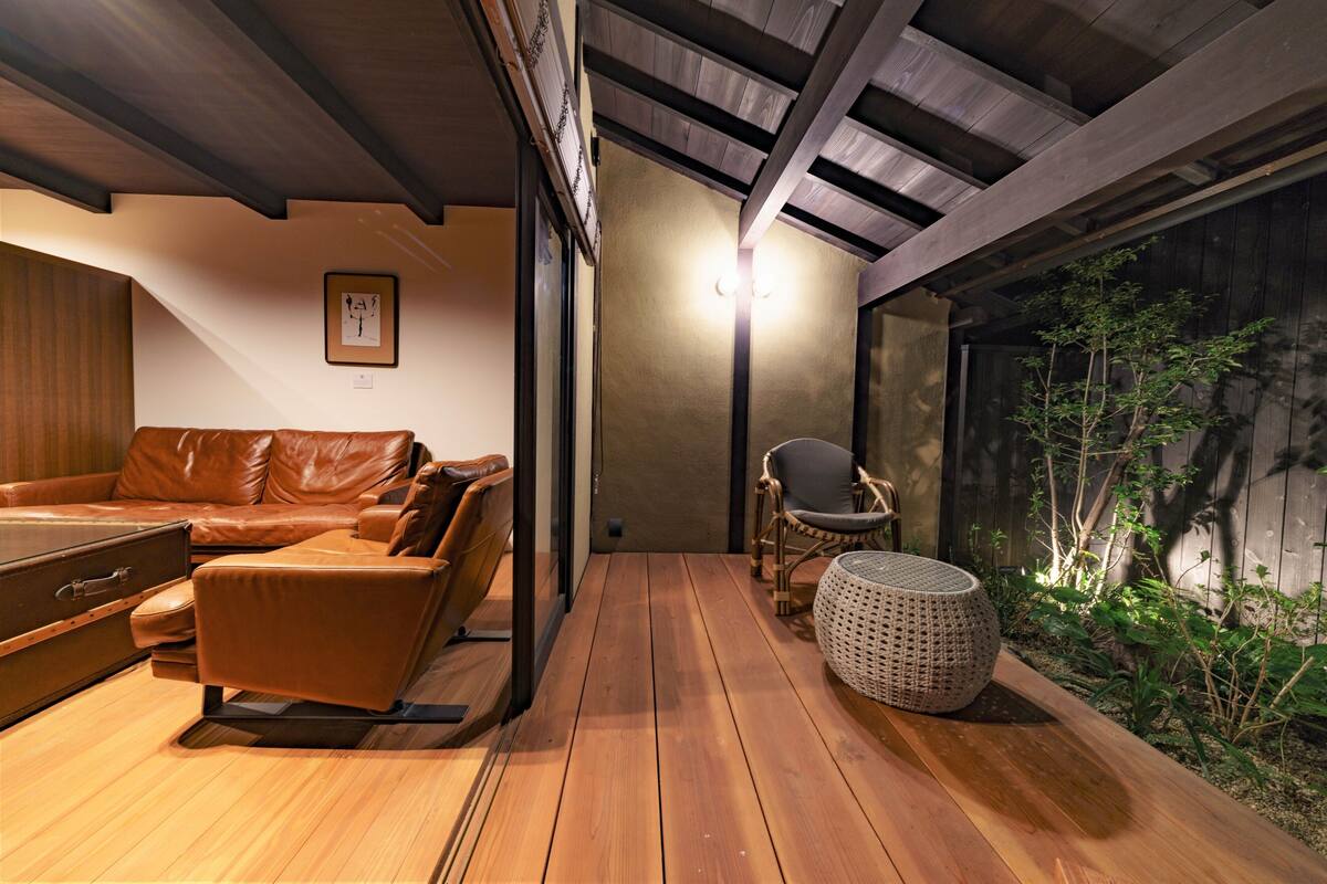 Puinen terassi, jossa on nahkasohva ja rottinkituoli, pieni pyöreä pöytä ja seinään kiinnitetty valaisin.