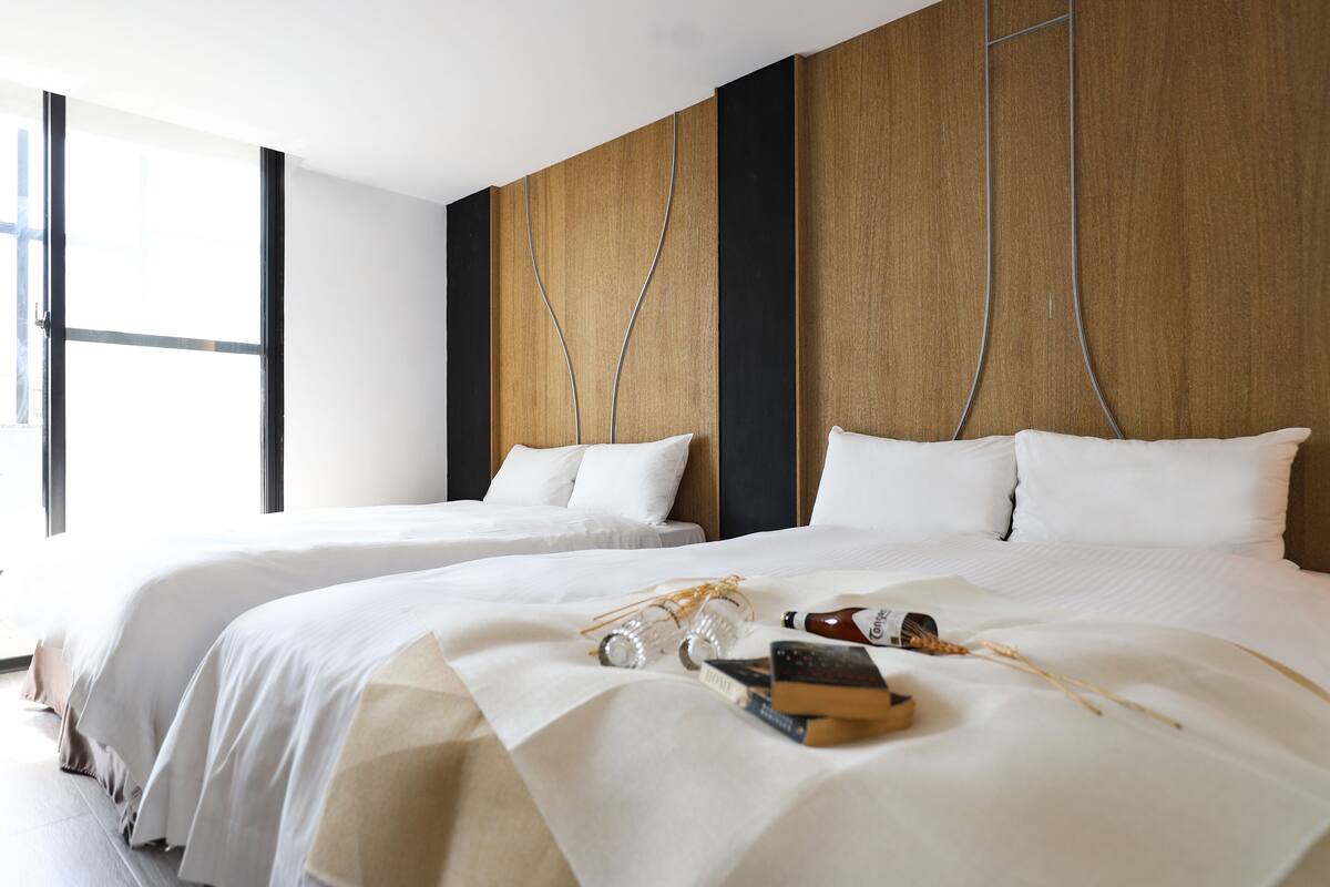 Quarto de hotel com duas camas, uma janela grande e uma cabeceira de madeira.