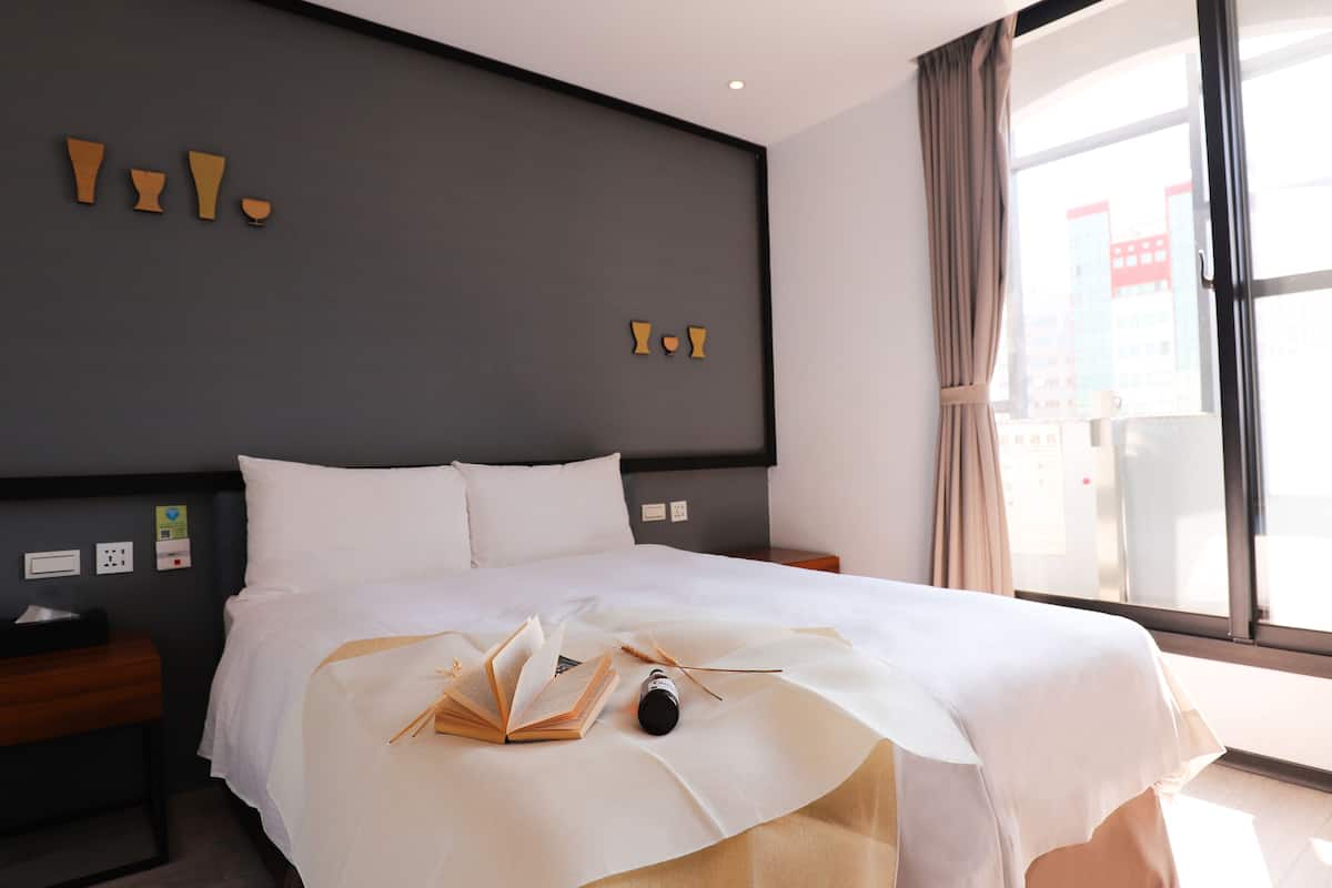 Quarto de hotel com uma cama grande, uma mesinha de cabeceira, cortinas numa janela e uma parede com formas geométricas.