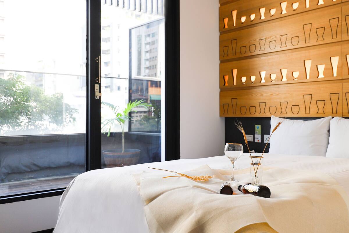 Quarto de hotel com uma cama grande, janela com vista para prédios e parede com formas recortadas.