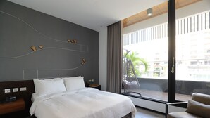A modern hotel room with a large bed, a wooden nightstand, a chair, and a balcony with a view of buildings and greenery. - Hotel Initial-Beer (Tainan)