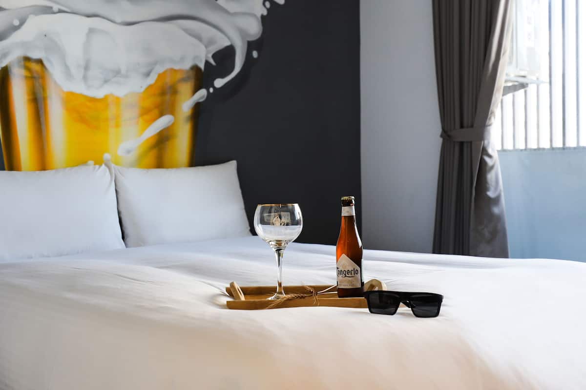 Cama bem arrumada com lençóis brancos, uma garrafa de cerveja Angerlo, uma taça de vinho e óculos de sol em um suporte.
