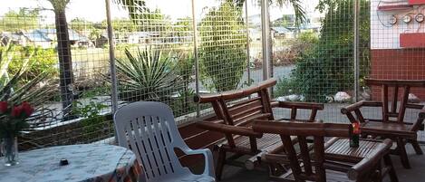 A covered outdoor seating area with wooden tables and chairs, a white plastic chair, and a table with a floral tablecloth.