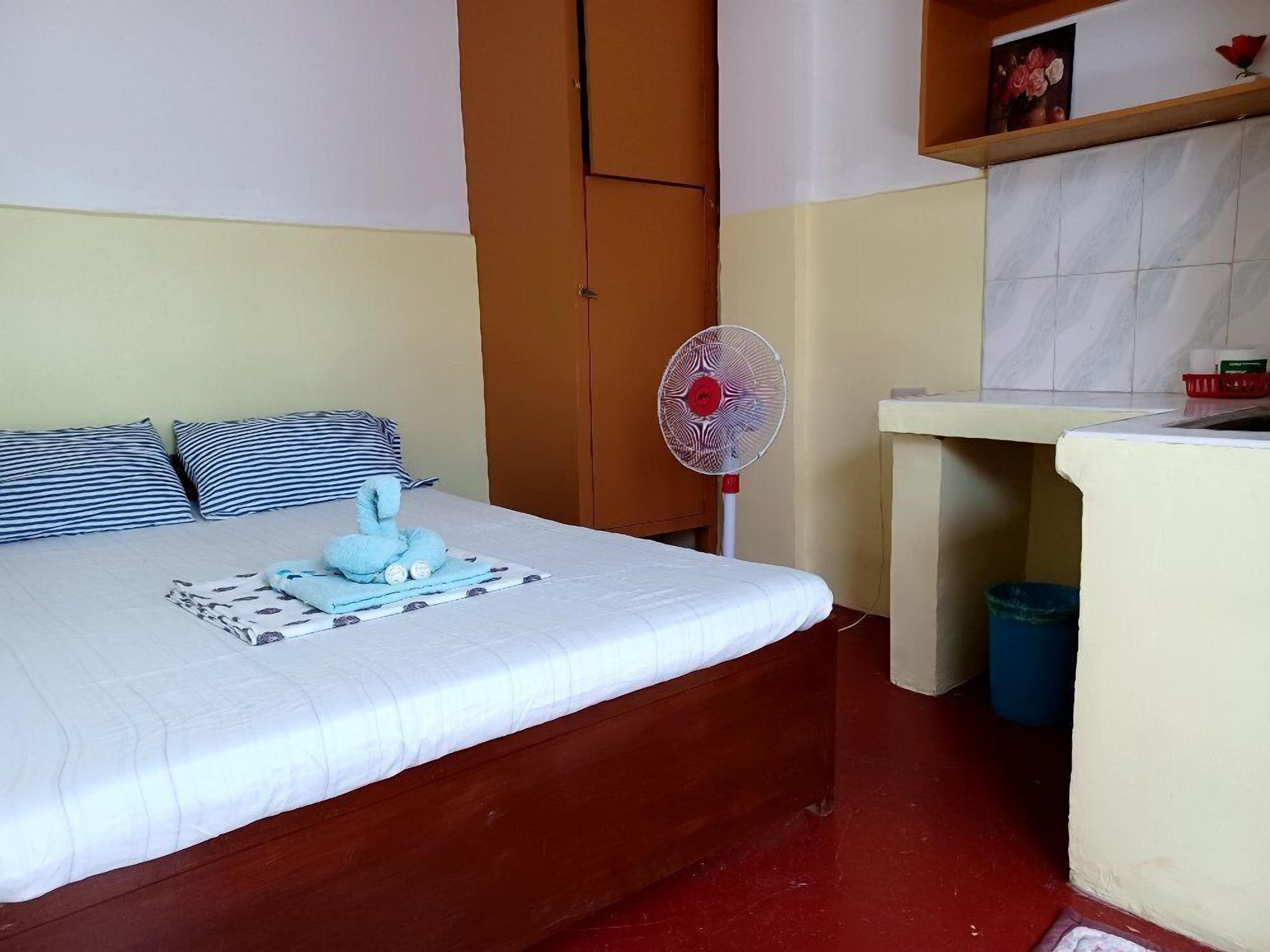 A bedroom with a bed, a standing fan, a wooden wardrobe, a small shelf with a picture, and a trash bin.