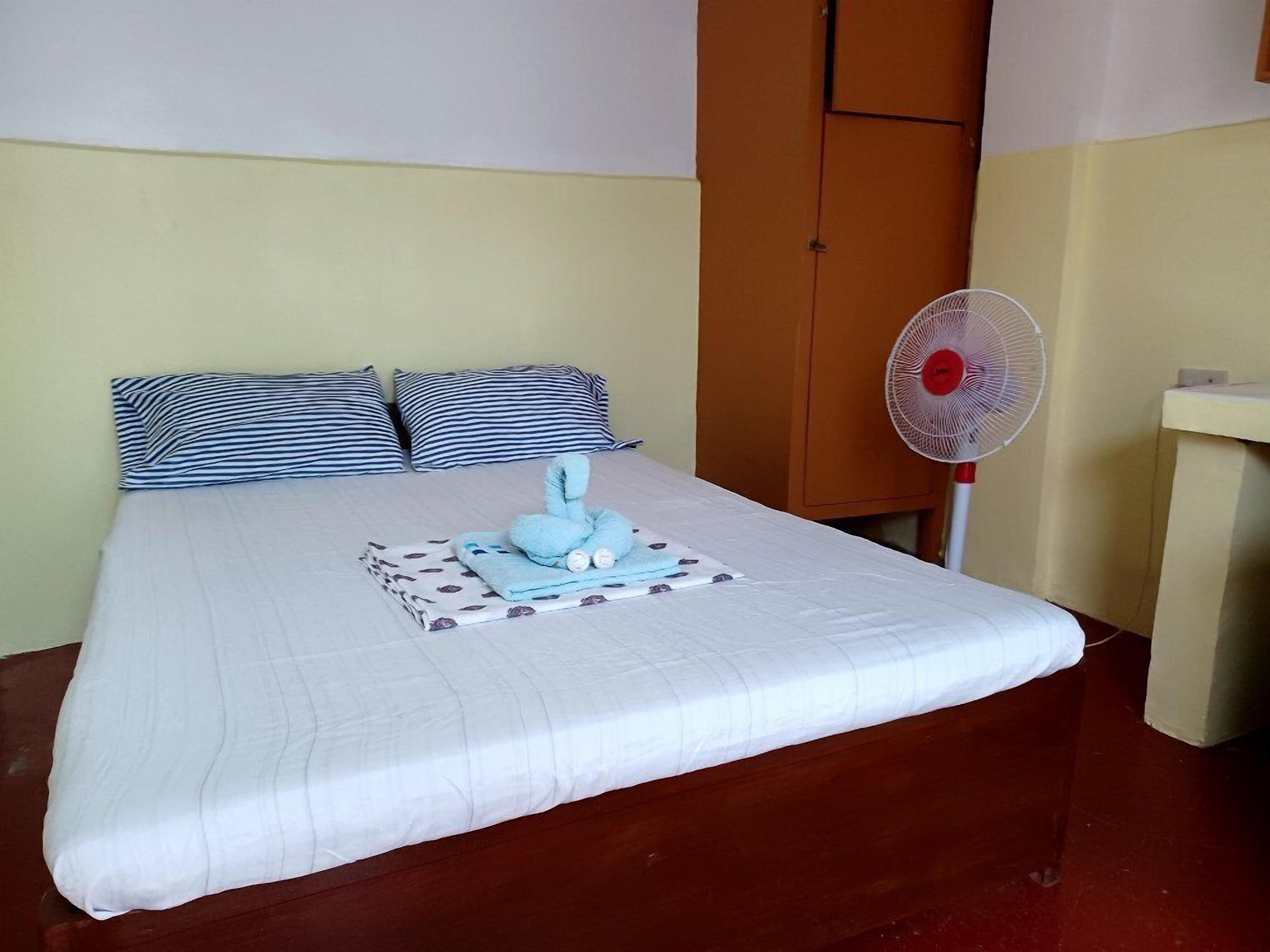 A neatly made bed with striped pillows, a white comforter, and a blue towel arrangement. A white standing fan is positioned to the right of the bed.